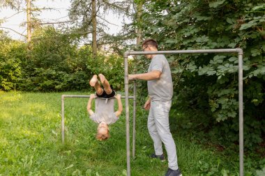 Baba ve çocuk, çocuk onu desteklerken çocuk baş aşağı sallanırken oyun alanında keyifli vakit geçiriyorlar. Yeşillik onları çevrelemiş, neşeli atmosferi geliştirmiş..