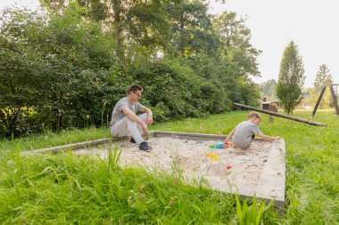 Bir baba, çocuğu oyuncaklarla oynarken kum havuzunun kenarında oturur ve neşeli bir an yaratır. Çevreleri yemyeşil ve ağaçlarla çevrili, açık havada sıcak bir günün tadını çıkarıyorlar..