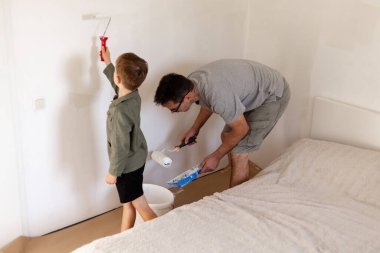 Genç oğlu ona yardım ederken bir adam yatak odasının duvarlarını boyuyor. Her ikisi de bağlarını güçlendiren ve odaya yeni bir görünüm kazandıran bir hafta sonu aktivitesinden zevk alıyor..