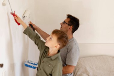 Bir baba ve küçük oğlu yatak odasının duvarlarını birlikte boyuyorlar. Çocuk boya fırçasıyla uğraşırken küçük bir silindir kullanıyor. Yeni ve temiz bir görünüm yaratıyorlar..