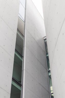 Büyük cam pencereli uzun beton yapılar modern bir şehir ortamı yaratır. Tasarım bulutlu bir gökyüzü altında keskin çizgiler ve minimalist özellikler sergiliyor.