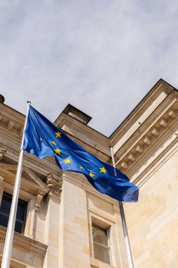 Avrupa Birliği bayrağı büyük tarihi bir binanın önündeki bayrak direğinde belirgin bir şekilde sergileniyor. Gökyüzü parçalı bulutlu, derinlik katıyor..