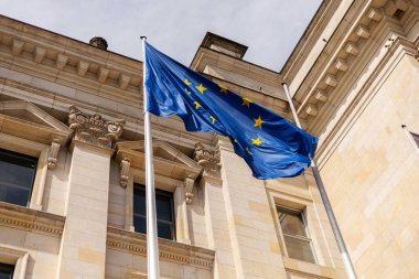 Canlı bir Avrupa Birliği bayrağı rüzgarda dalgalanıyor, güneşli bir günde klasik bir mimari arka plana karşı yıldızlarını ve mavi rengini gösteriyor.