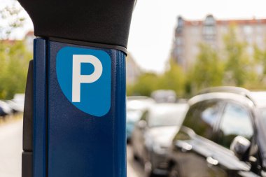 Parlak, güneşli bir günde kalabalık bir şehir ortamında park halindeki birkaç arabanın yanında bir parkmetre görünür. Arka planda ağaçlar ve binalar görülebilir..