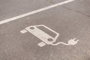 Elektrikli taşıt şarj istasyonunu temsil eden açık bir sembol bir park alanının kaldırımına resmedilmiştir. Sahne doğal gün ışığıyla aydınlandı..