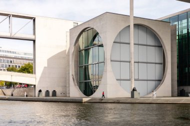 Berlin, Almanya - 1 Eylül 2025: Berlin 'de, insanlar Eylül ayında parçalı bulutlu bir gökyüzünün altında geniş dairesel pencerelere sahip, deniz kenarında çarpıcı bir modern binanın önünden geçtiler..