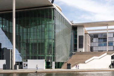 Berlin, Almanya - 1 Eylül 2025: Ziyaretçiler Berlin 'deki modern bir binanın parlak cam yapısının yanından geçtiler. Gün ortası güneşi nehir kıyısı boyunca mimariye ilginç gölgeler bırakıyor..
