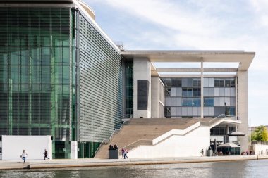 Berlin, Almanya - 1 Eylül 2025: İnsanlar Berlin 'de deniz kenarındaki etkileyici modern binaların önünden geçiyorlar. Temiz çizgiler ve cam cephe, şehrin güneşli bir günde yaptığı mimari yeniliğin altını çiziyor..