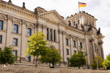 Berlin, Almanya - 1 Eylül 2025: Reichstag Binası, karmaşık mimari ve dalgalı bir Alman bayrağının yer aldığı Berlin 'de gururla duruyor. Ziyaretçiler çevredeki yeşilliğin tadını çıkarıyorlar.