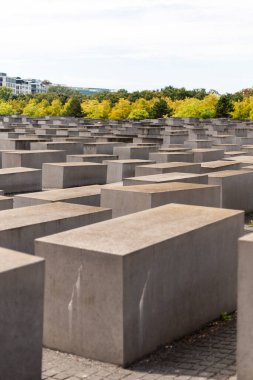 Berlin, Almanya - 1 Eylül 2025 Berlin 'de ziyaretçiler, güneşli bir günde canlı ağaçlar ve mavi gökyüzü ile çevrili Yahudi Soykırımı anıtının büyük gri blokları arasında yürürler.