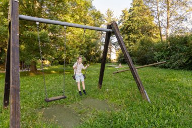İki çocuk güneşli bir parkta mutlu bir şekilde oynuyorlar. Bir çocuk salıncakta sallanıyor, gülümsüyor ve temiz havanın tadını çıkarırken yeşil ağaçlar ve çimenler etrafını sarıyor..
