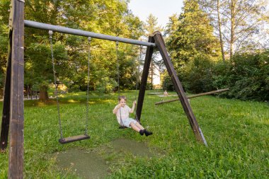 İki çocuk yemyeşil ağaçlarla dolu bir parkta neşeyle oynuyorlar. Bir çocuk salıncakta sallanırken diğeri yakınlardaki çimleri keşfediyor..