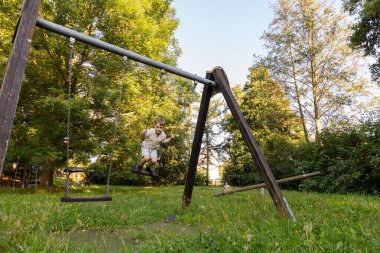 İki çocuk yeşil çimenler ve uzun ağaçlarla dolu geniş bir parkta salıncaklarda neşeyle oynuyorlar. Güneş parlıyor, sıcak ve neşeli bir atmosfer yaratıyor..