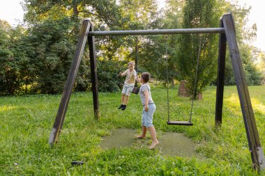 İki çocuk canlı bir parkta salıncakta sallanırken mutlu. Sahne yemyeşil çimenler ve ağaçlarla dolu, neşeli bir yaz havası yayıyor..