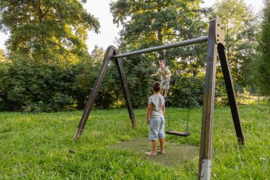 İki çocuk parkta salıncakta eğleniyor. Bir çocuk mutlu bir şekilde sallanırken diğeri ağaçlar ve otlarla çevrili sıcak ve güneşli günün tadını çıkarıyor..