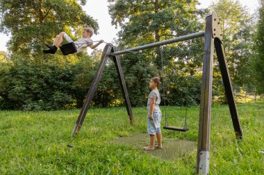 İki çocuk yeşil bir parkta neşeyle oynuyorlar. Bir çocuk yüksekte sallanırken diğeri heyecanla izliyor. Parlak gün ışığı ağaçların arasından süzülüp, canlı bir sahne yaratıyor..