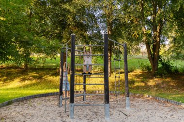 İki çocuk geniş bir parktaki oyun parkına tırmanırken eğleniyorlar. Güneş ışığı ağaçların arasından süzülür ve birlikte oynarken neşeli bir atmosfer yaratır..