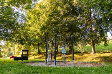 İki çocuk güneşli bir parktaki tırmanma parkında mutlu mesut oynuyorlar. Parlak yeşil ağaçlar oyun alanının etrafını sarıp, dışarıda eğlenmek için neşeli ve canlı bir atmosfer yaratıyor..