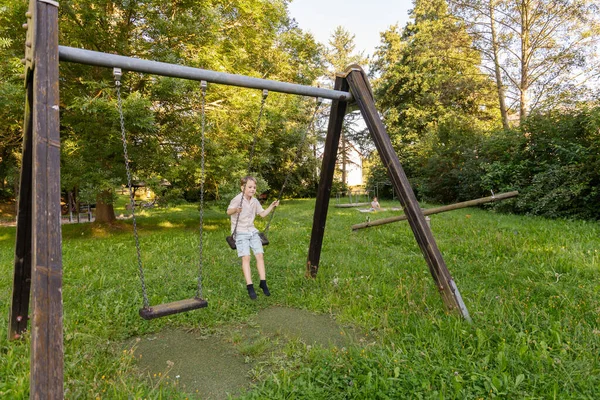 İki çocuk güneşli bir parkta mutlu bir şekilde oynuyorlar. Bir çocuk salıncakta sallanıyor, gülümsüyor ve temiz havanın tadını çıkarırken yeşil ağaçlar ve çimenler etrafını sarıyor..