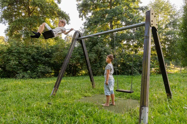 İki çocuk yeşil bir parkta neşeyle oynuyorlar. Bir çocuk yüksekte sallanırken diğeri heyecanla izliyor. Parlak gün ışığı ağaçların arasından süzülüp, canlı bir sahne yaratıyor..
