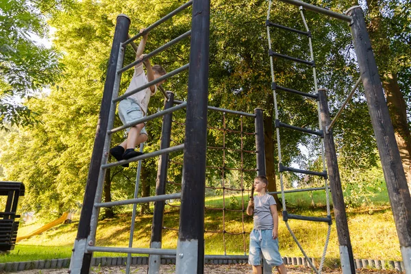 İki çocuk oyun alanında eğleniyor, biri metal yapılara tırmanırken diğeri izliyor. Sahne aydınlık ve neşeli, açık hava aktivitelerini vurguluyor..
