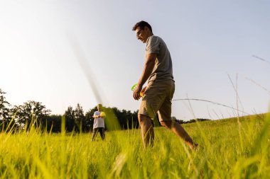 İki aile ferdi dışarıda keyifli bir anı paylaşıyor. Baba ve oğul güneşli bir günde açık mavi gökyüzünün altında yemyeşil bir tarlada eğlenceli aktivitelere girişiyor..