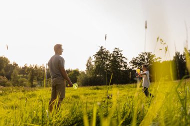 Baba ve oğul, uzun çimenler ve ağaçlarla çevrili yemyeşil bir tarlada eğlenceli bir oyun oynuyorlar. Güneş batarken güzel vakit geçiriyorlar..