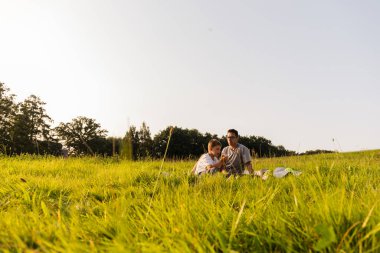 Baba ve oğul yemyeşil bir çayırda battaniyeye oturup sıcak güneşin altında hikayeler ve kahkahalar paylaşarak doğada kalıcı anılar yaratırlar..
