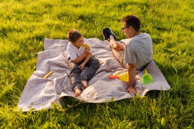 Bir baba ve küçük oğlu yemyeşil bir parkta battaniyenin üzerinde dinleniyorlar. Atıştırmalıkları paylaşırlar ve renkli oyuncaklarla oynarlar, güneşli bir öğleden sonranın tadını çıkarırlar. Kahkahalar havayı doldurur.