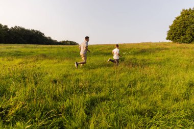 Bir baba ve oğlu, yemyeşil bir arazide koşuşturarak dışarıda kaliteli zaman geçirmekten zevk alır. Güneş ışığı bu güzel öğleden sonra sıcak bir atmosfer yaratır..