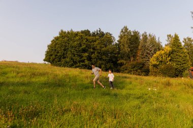 Bir çocuk ve babası yemyeşil bir arazide neşeyle koşuyorlar. Güneş parlıyor ve ağaçlar güzel bir zemin sağlıyor. Açık havada şakacı bir bağ kuruyorlar..