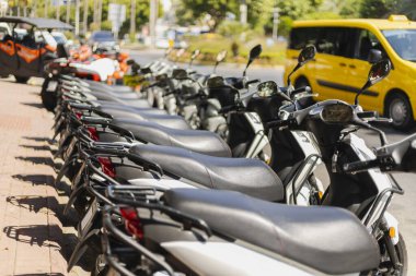 Bir sıra park etmiş scooterlar işlek bir şehir caddesinde. Güneş ışığı olay yerinde parlarken sarı bir taksi yakınlarda bekliyor. Atmosfer canlı ve şehirli hissettiriyor..