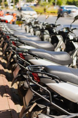 Bir sıra park etmiş scooter 'lar kalabalık bir cadde boyunca binicileri bekliyor. Güneş parıldıyor, parlak tasarımı ve araçların canlı renklerini vurguluyor..