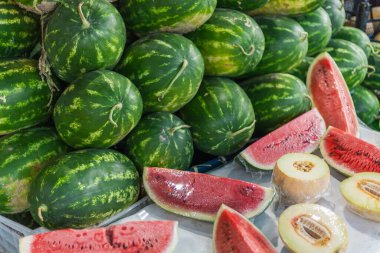 Parlak yeşil karpuzlar yüksek istiflenirken sulu karpuz dilimleri ve bütün bir kavun masaya oturup yaz meyvelerinin bolluğunu gözler önüne seriyor..