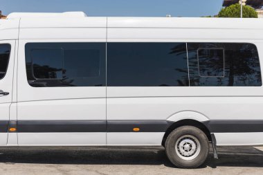 Parlak güneşli bir günde etrafı ağaçlar ve binalarla çevrili asfalt bir zemine park edilmiş büyük beyaz bir yolcu minibüsü var. Minibüste renkli camlar ve parlak bir tasarım yer alıyor..