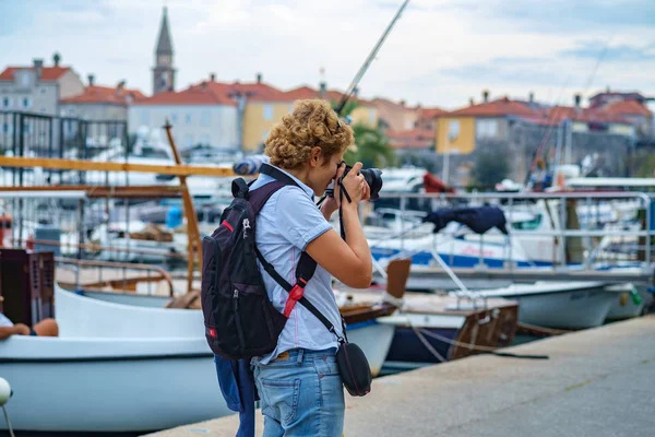 Photographer with a backpack taking travel photography. Woman photographer shooting with dslr camera walking along the promenade with beautiful maritime landscape in backgroundGirl from behind photographing on the seafront promenade