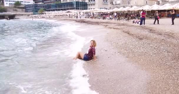 Mladá hezká blondýnka dívka v košili a krátké kalhoty na pláži u moře představuje pro fotografa. Její šaty jsou mokré po vlně již pokryl ji