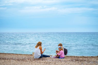 Budva, Karadağ, 06 Ekim 2018: İki çocuk - bir kız ve bir erkek - genç güzel sarışın kızla sahilde denizin kenarında piknik vardır. Aile hayran sakin mavi deniz, kum üzerine oturan bir kadın denizin üstünde onun smartphone çekiyor