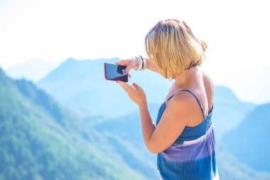 Genç güzel sarışın turist onun smartphone, yeşil ormanıyla kaplı dağları manzara resimlerini alır. Parlak güneşli bir günde
