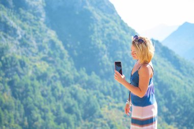Genç güzel sarışın turist onun smartphone, yeşil ormanıyla kaplı dağları manzara resimlerini alır. Parlak güneşli bir günde