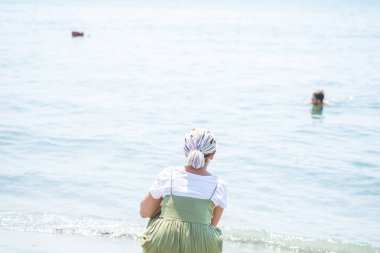 Genç kadın bir hafif pamuklu sundress çok renkli dreadlocks ile plaj su kenarında squats ve denizde görünüyor. Arkadan görüntülemek