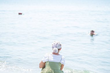 Genç kadın bir hafif pamuklu sundress çok renkli dreadlocks ile plaj su kenarında squats ve denizde görünüyor. Arkadan görüntülemek
