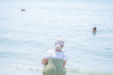 Genç kadın bir hafif pamuklu sundress çok renkli dreadlocks ile plaj su kenarında squats ve denizde görünüyor. Arkadan görüntülemek