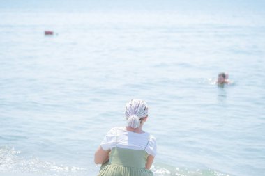 Genç kadın bir hafif pamuklu sundress çok renkli dreadlocks ile plaj su kenarında squats ve denizde görünüyor. Arkadan görüntülemek