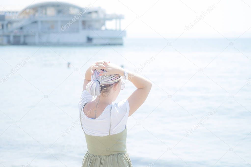 Young woman with multi-colored dreadlocks in a light cotton sundress squats on the beach at the water's edge and looks at the sea. View from the back
