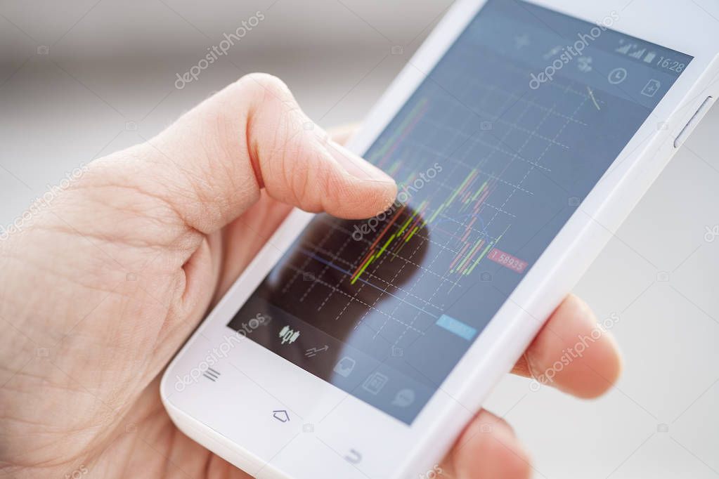 Close up finger hand of a trader touching smartphone screen with trading interface dashboard with candlestick chart in mobile trading forex or stock application. Forex and investment business technology concept