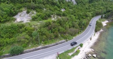 Quadcopter deniz kıyısı boyunca uzanan dar bir asfalt yol üzerinde uçuyor. Yolun diğer tarafında yeşil ağaçlarla kaplı turkuaz denizi ve sahili görebiliyoruz. Arabalar, kamyonlar ve turist otobüsleri yol boyunca sürüyor