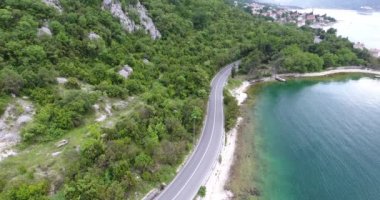 Quadcopter deniz kıyısı boyunca uzanan dar bir asfalt yol üzerinde uçuyor. Yolun diğer tarafında yeşil ağaçlarla kaplı turkuaz denizi ve sahili görebiliyoruz. Arabalar, kamyonlar ve turist otobüsleri yol boyunca sürüyor