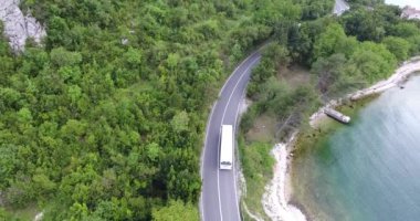 Quadcopter deniz kıyısı boyunca uzanan dar bir asfalt yol üzerinde uçuyor. Yolun diğer tarafında yeşil ağaçlarla kaplı turkuaz denizi ve sahili görebiliyoruz. Arabalar, kamyonlar ve turist otobüsleri yol boyunca sürüyor