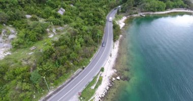 Quadcopter deniz kıyısı boyunca uzanan dar bir asfalt yol üzerinde uçuyor. Yolun diğer tarafında yeşil ağaçlarla kaplı turkuaz denizi ve sahili görebiliyoruz. Arabalar, kamyonlar ve turist otobüsleri yol boyunca sürüyor